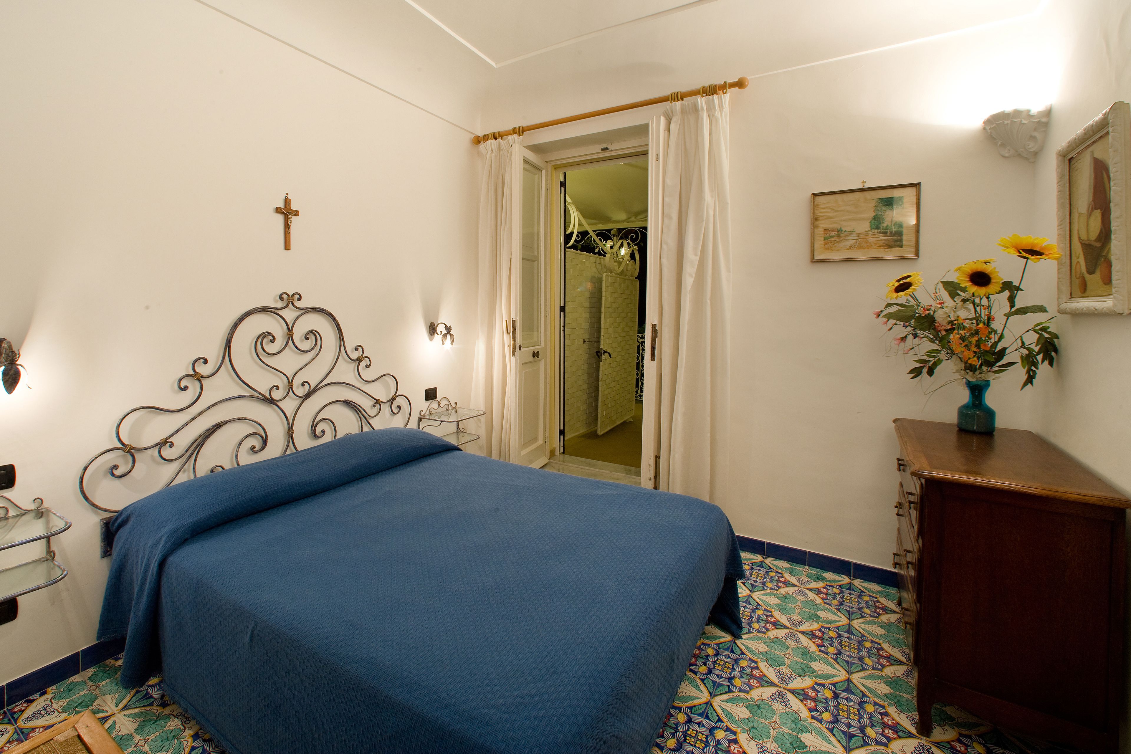 Un dormitorio con una cama, un aparador, un espejo, una cruz en la pared y una puerta que da a otra habitación.
