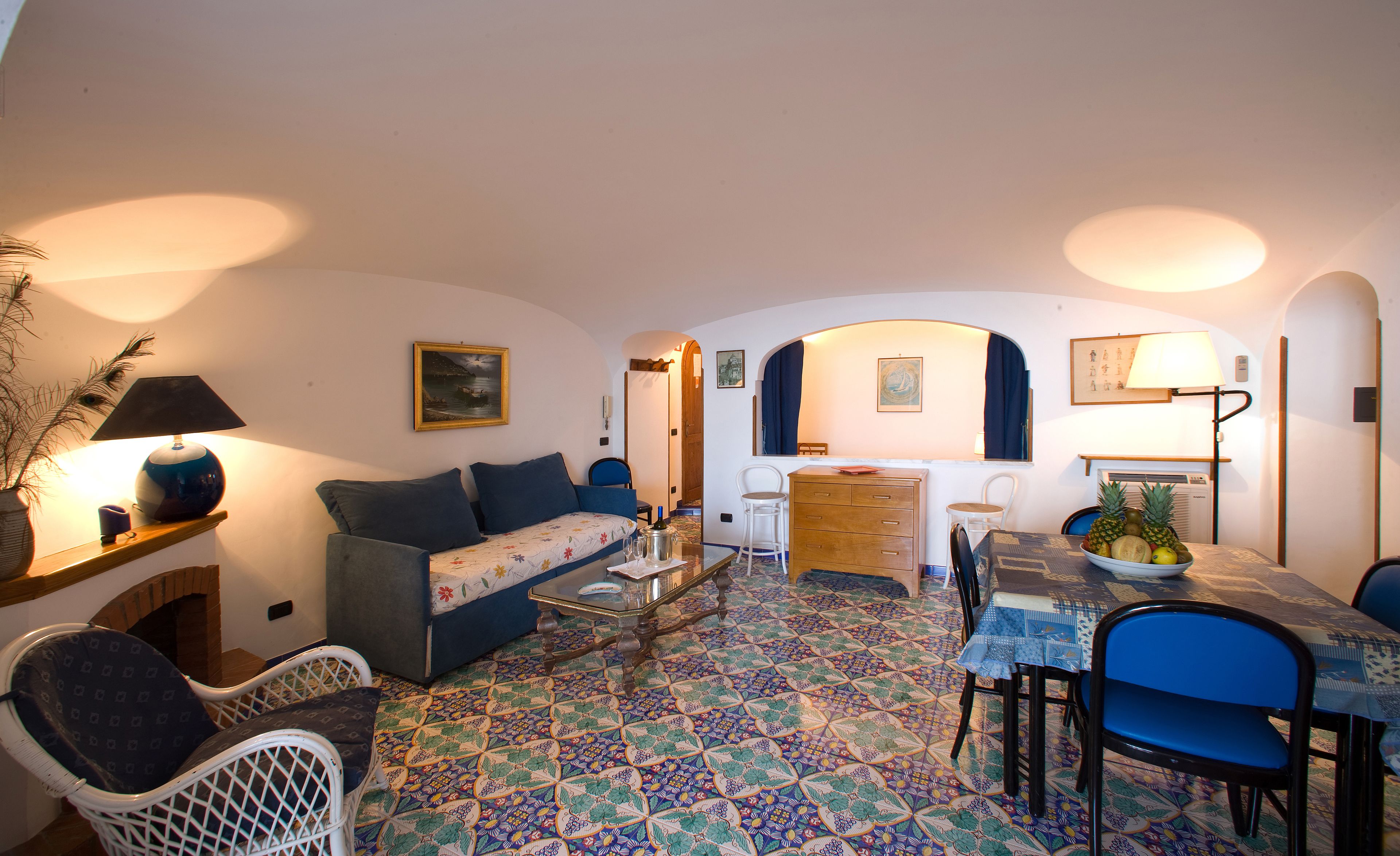 Ein gemütliches Wohnzimmer mit einer Couch, Sesseln, einem Couchtisch und einem Esstisch mit Stühlen. Der Boden ist gemustert, eine Lampe steht dort und es hängt ein Gemälde an der Wand.