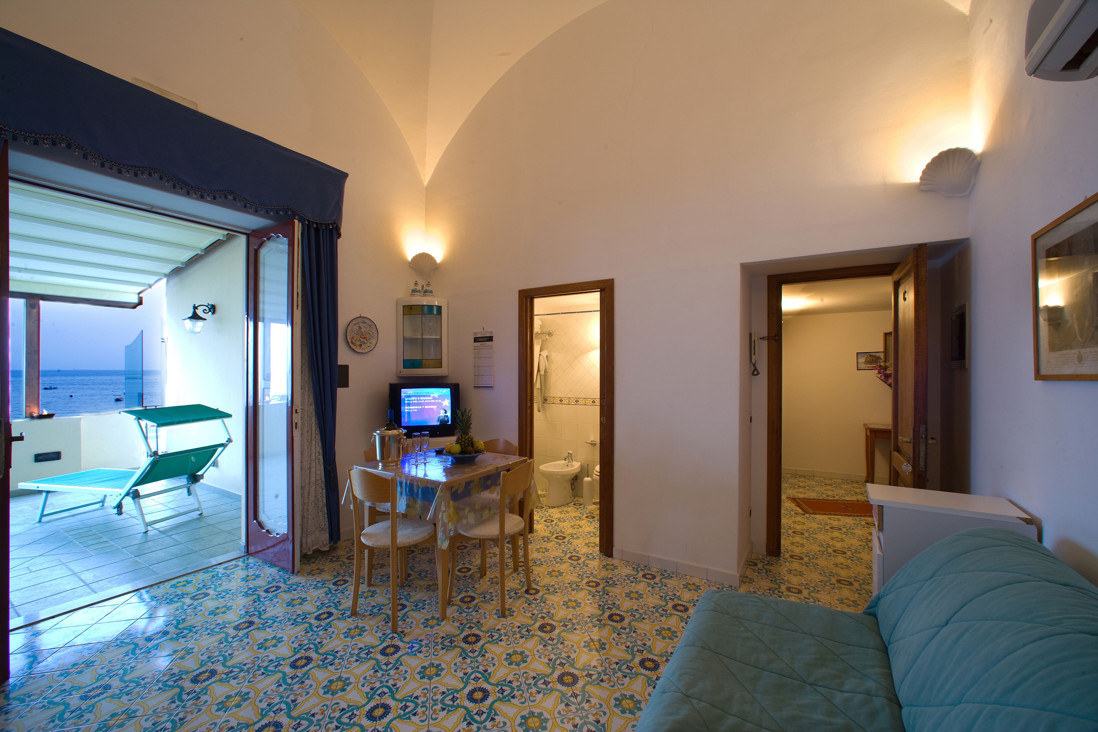 Una habitación con suelo de baldosas, una cama, una mesa con sillas, un televisor y vista al mar a través de una puerta abierta.