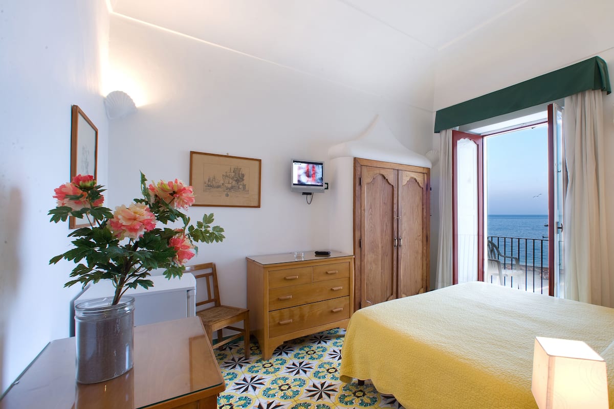 A bedroom with a bed, a wooden dresser, a desk with a vase of flowers, a television, and a view of the sea through an open door.