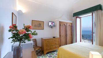 Un dormitorio con cama, cómoda de madera, escritorio con un jarrón de flores, televisión y vista al mar a través de una puerta abierta.