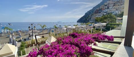 A balcony with purple flowers, a view of the beach, and a mountain in the background.