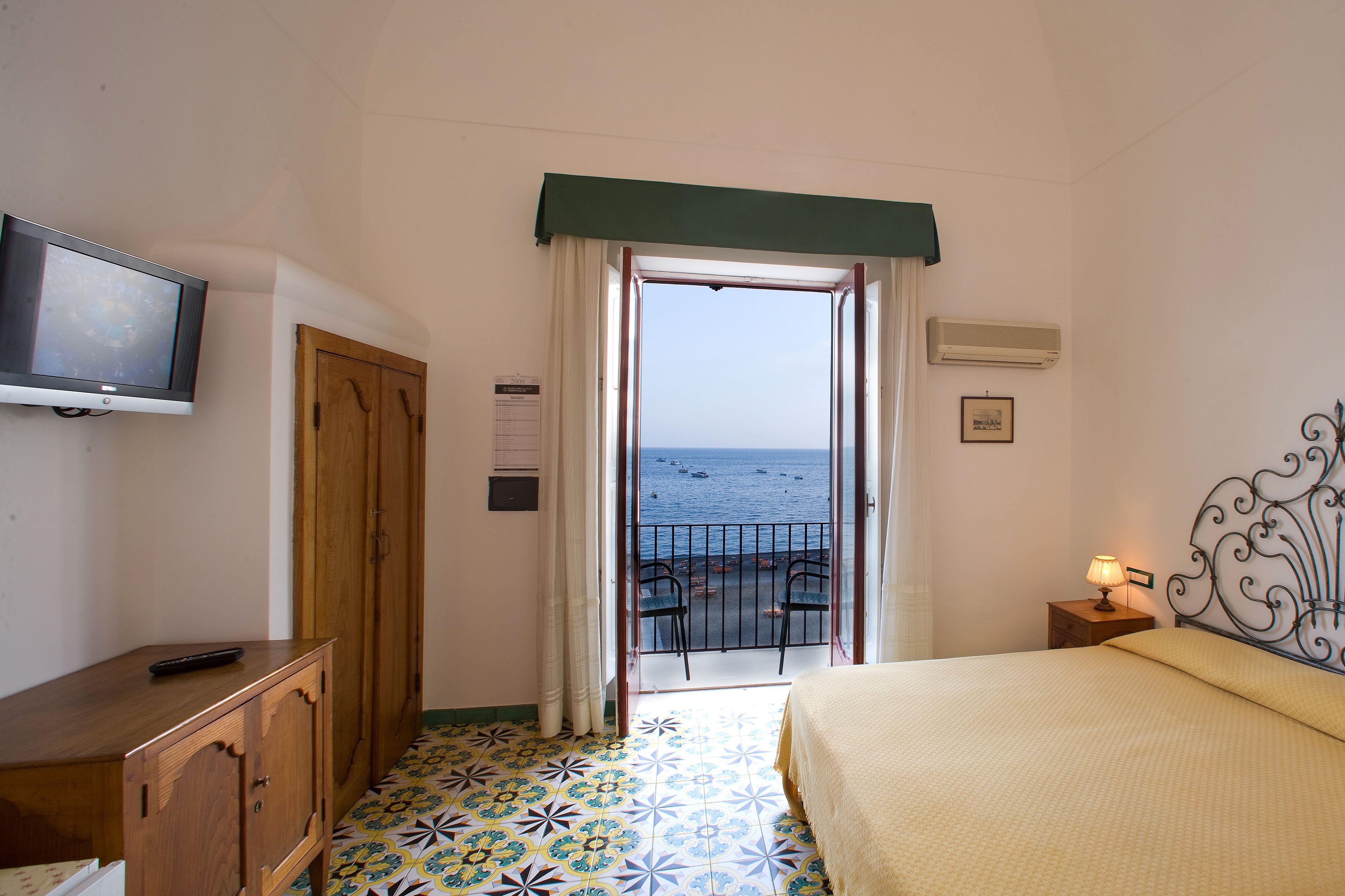 A bedroom with a bed, a TV, a wooden wardrobe, a small table with a lamp, and a balcony with a view of the sea.