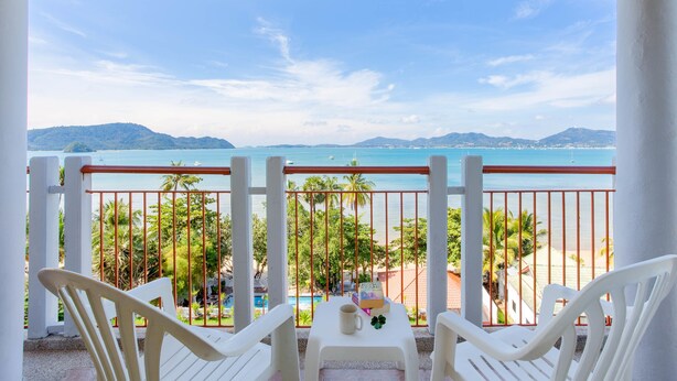 Un balcon avec des chaises blanches et une table, d’où l’on peut admirer une plage et une mer turquoise.