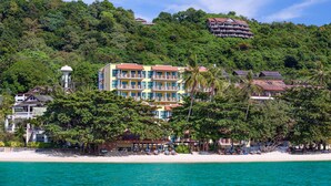 Un hôtel en bord de mer, avec plusieurs étages, une plage en premier plan et une végétation luxuriante sur la colline.