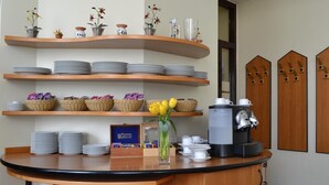 A hotel reception area with a curved counter, stacks of plates, baskets of snacks, a coffee machine, and decorative shelves with flowers and vases. - Fair Hotel am Rathaus (Schwalbach am Taunus)