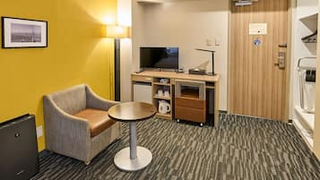 Habitación de hotel con un sofá individual, una pequeña mesa redonda, televisión, minibar y una puerta de madera.