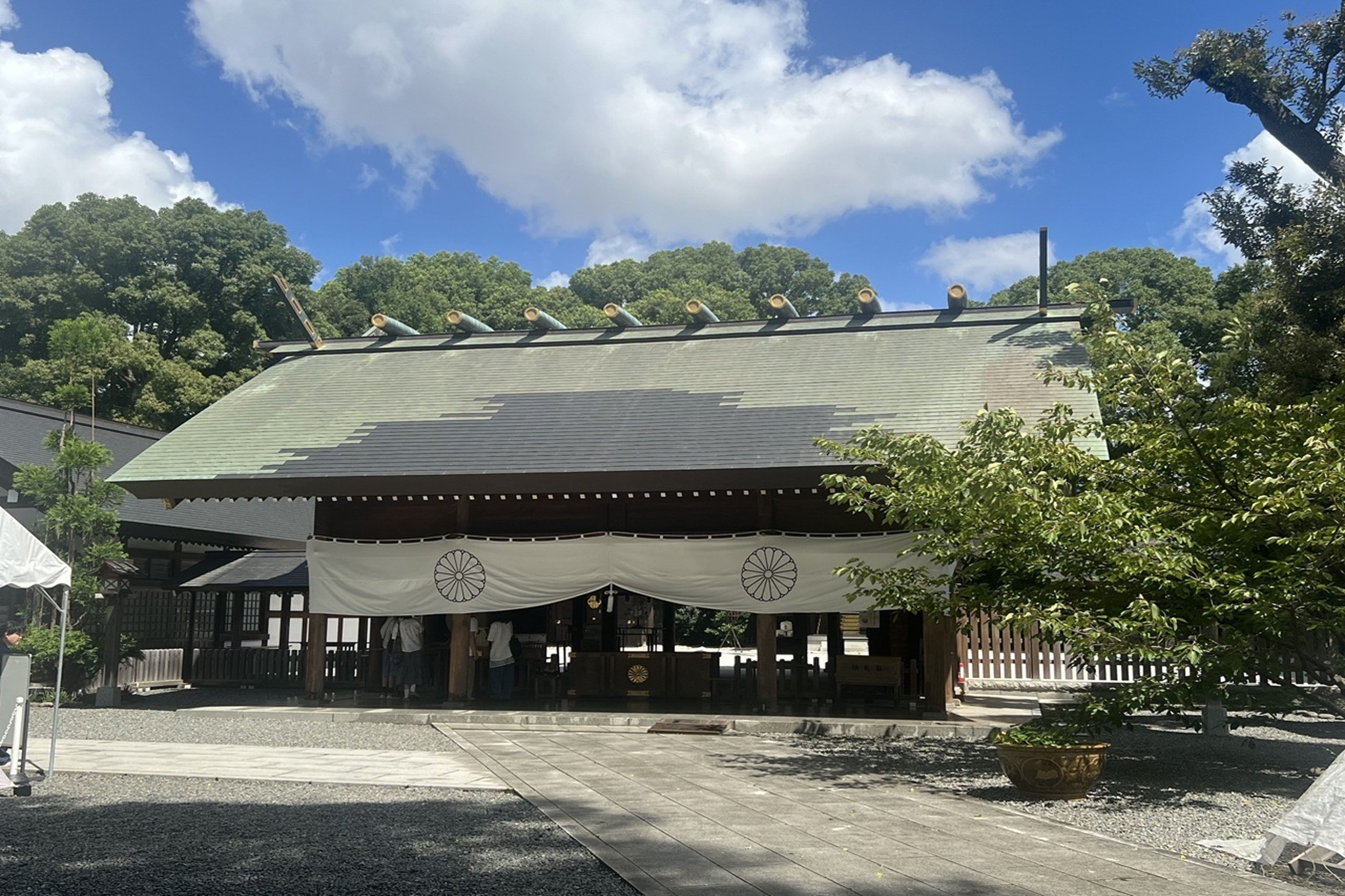 A traditional building with a tiled roof, a covered entrance, and a garden area with a tree and a bench.