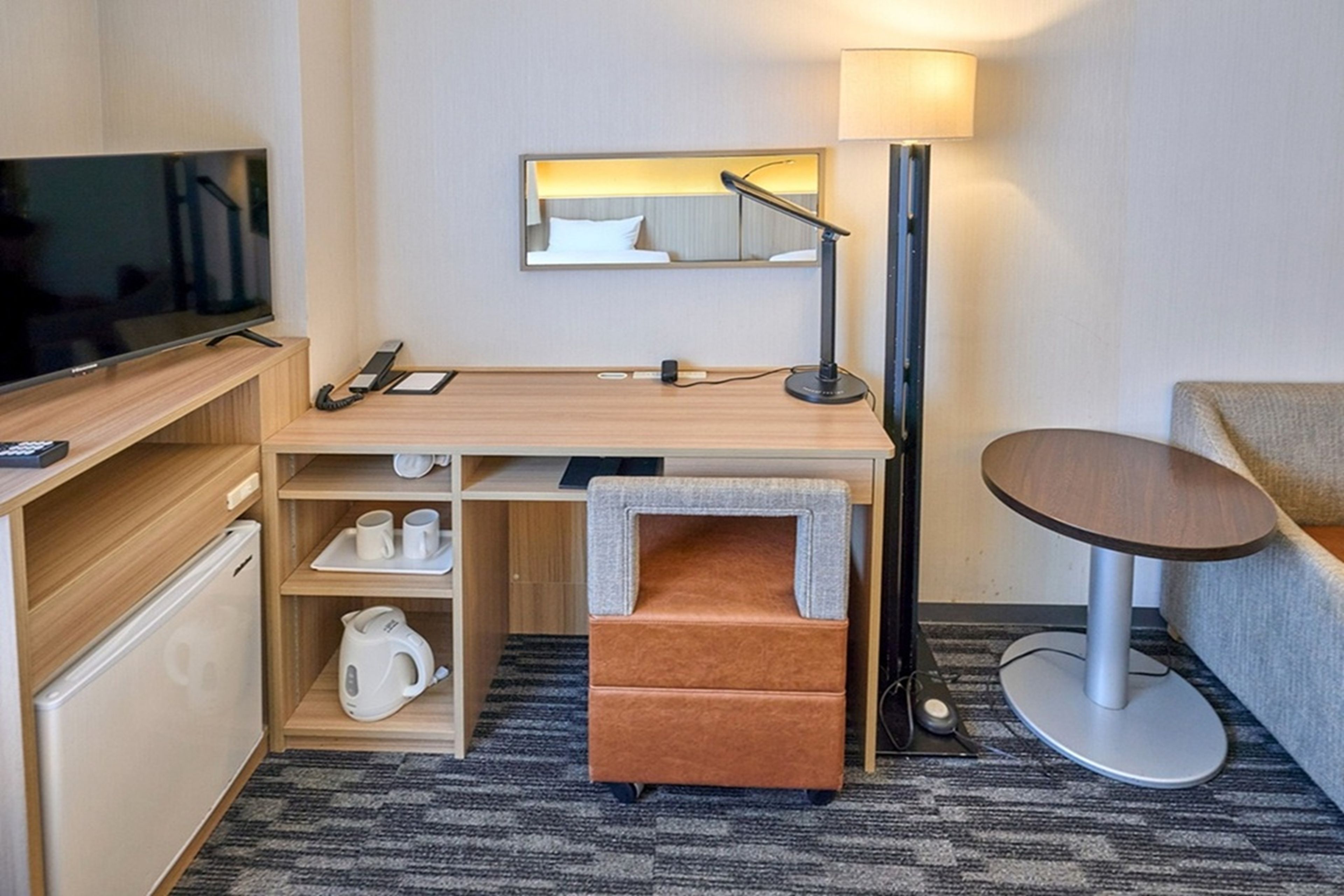 A compact hotel room with a flat-screen TV, a desk with a lamp, a small sofa, and a coffee maker.