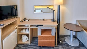 Habitación de hotel compacta con televisor de pantalla plana, escritorio con lámpara, un pequeño sofá y cafetera.