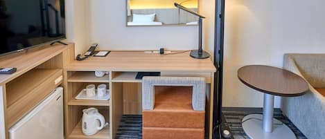 A compact hotel room with a flat-screen TV, a desk with a lamp, a small sofa, and a coffee maker.