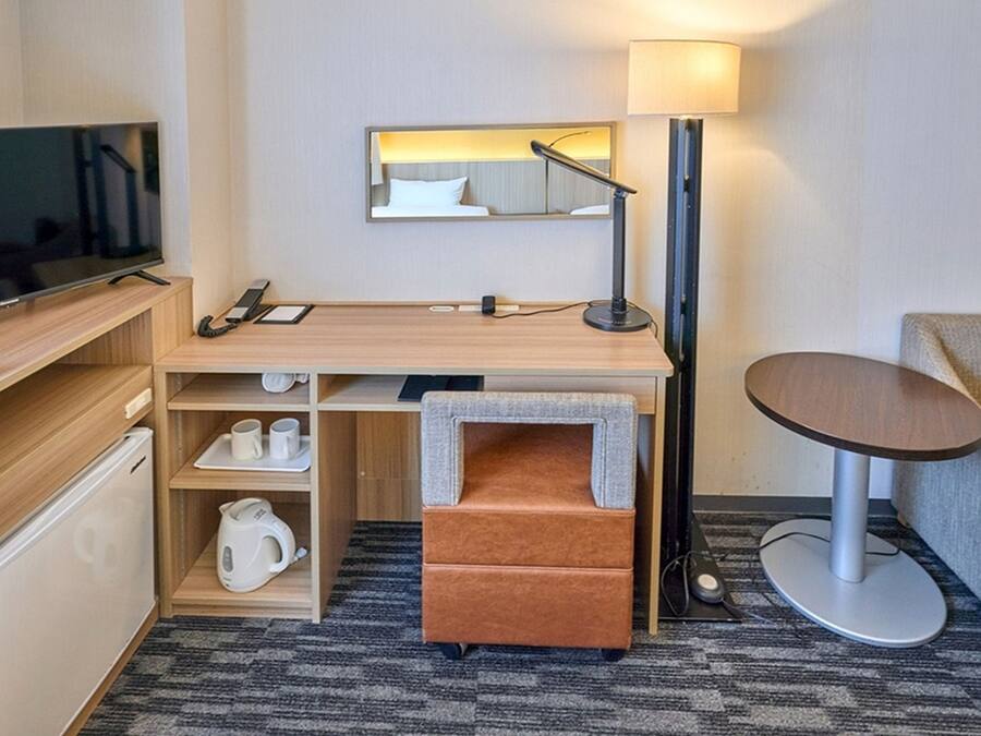 Habitación de hotel compacta con televisor de pantalla plana, escritorio con lámpara, un pequeño sofá y cafetera.