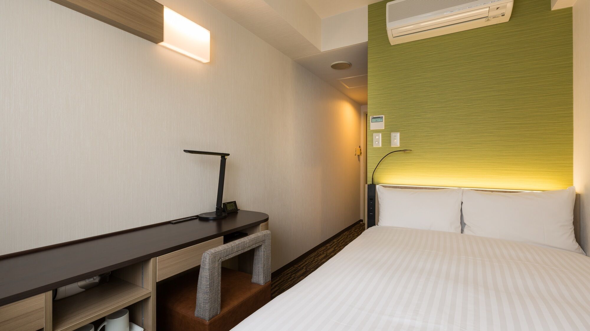 A hotel room with a bed, a desk with a chair, a standing lamp, and an air conditioning unit.