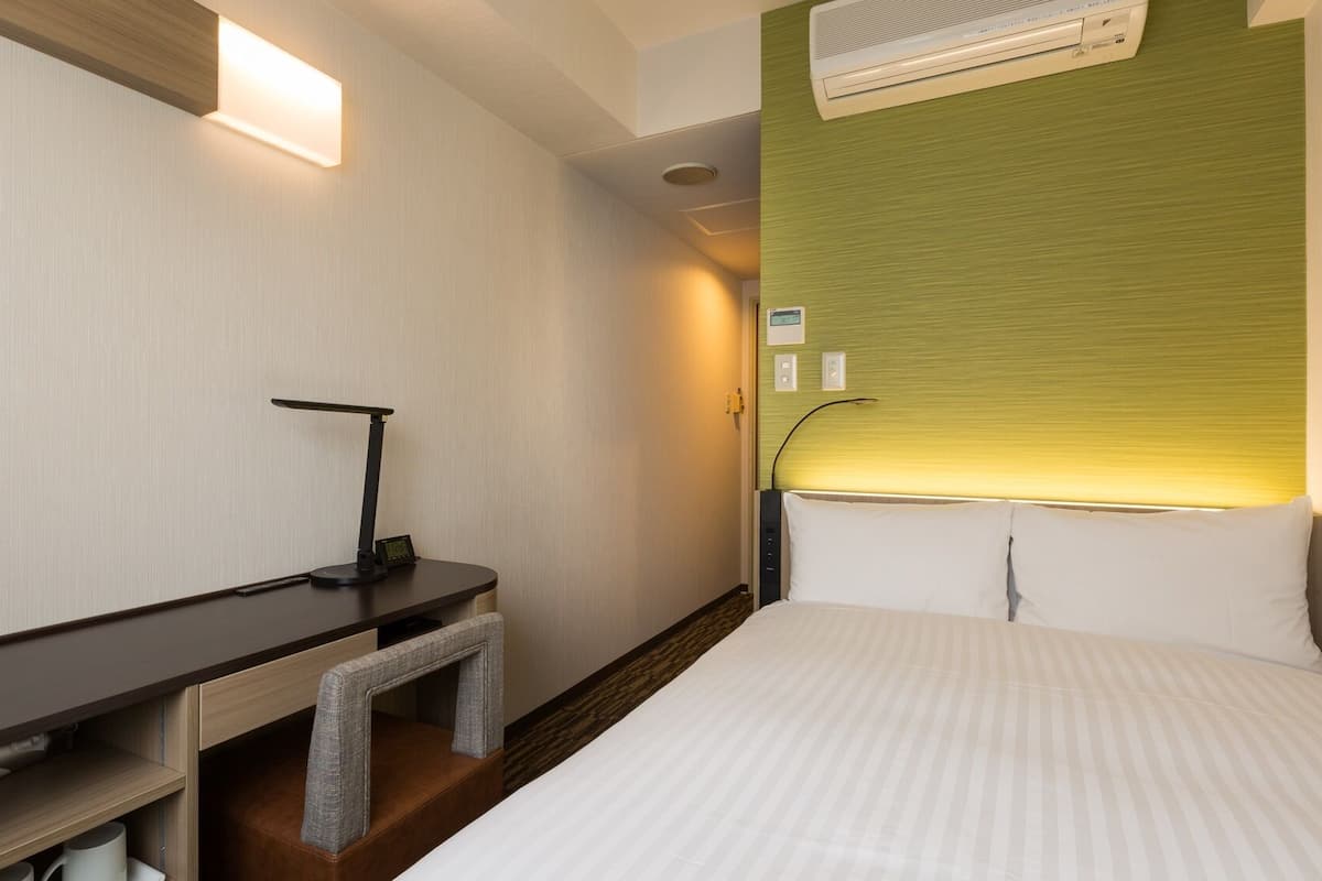 A hotel room with a bed, a desk with a chair, a standing lamp, and an air conditioning unit.