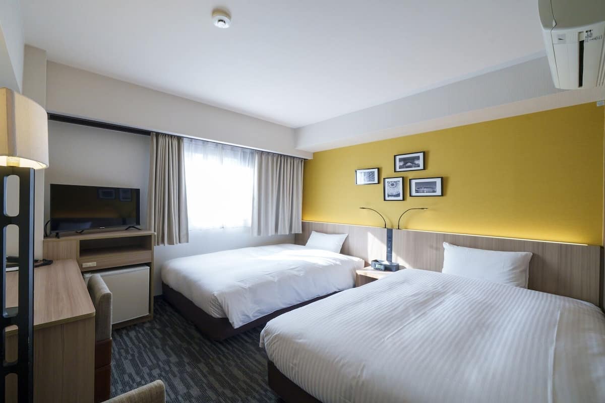 A hotel room with two beds, a desk with a TV, a lamp, and a wall-mounted air conditioner.