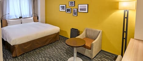 Quarto de hotel com uma cama grande, uma poltrona pequena, uma mesa redonda e uma parede com quadros.
