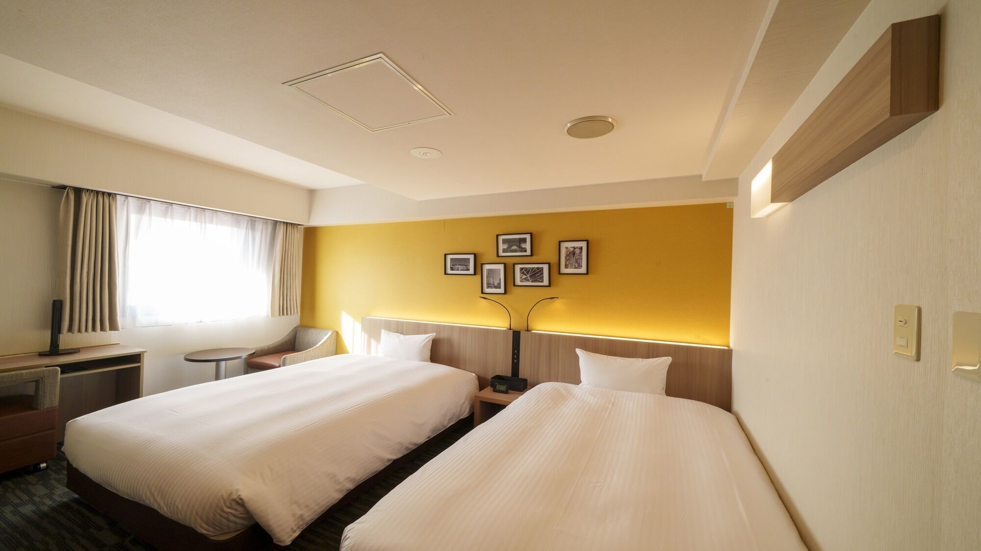 A hotel room with two beds, a desk, a chair, a small table, and a wall with framed pictures.