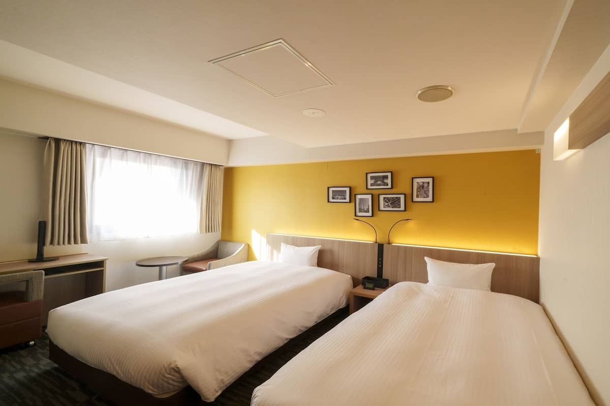 A hotel room with two beds, a desk, a chair, a small table, and a wall with framed pictures.