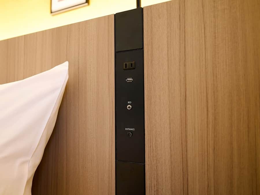 Ein Hotelzimmer mit einem hölzernen Kopfteil, einem weißen Kissen und einer Bedieneinheit mit Knopf für die Bettenverstellung und Eingangstaste.