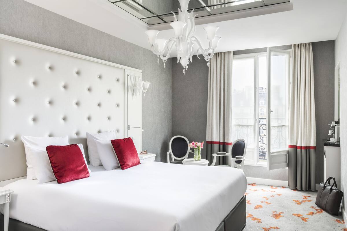 A hotel room with a large bed, a bedside table, a chair, a desk, a mirror, a chandelier, and a view of the city through the window.