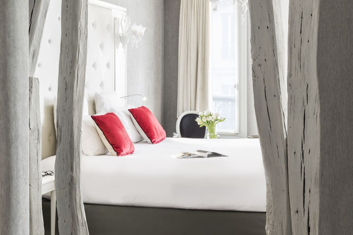 A neatly made bed with white and red pillows, a black chair, and a vase with white flowers.