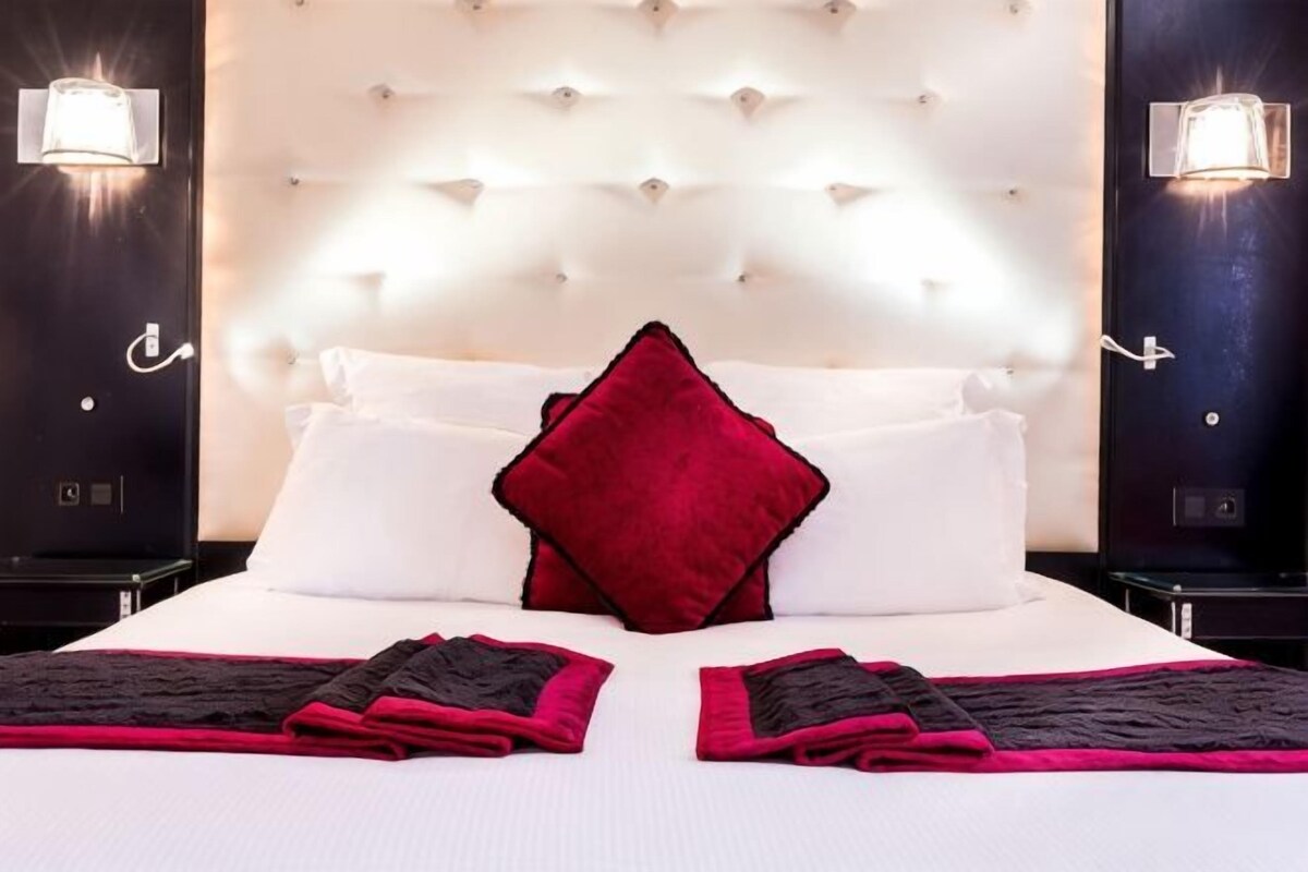 Habitación de hotel con cabecera capitonada, ropa de cama blanca, un cojín rojo decorativo y dos mesitas de noche con lámparas.