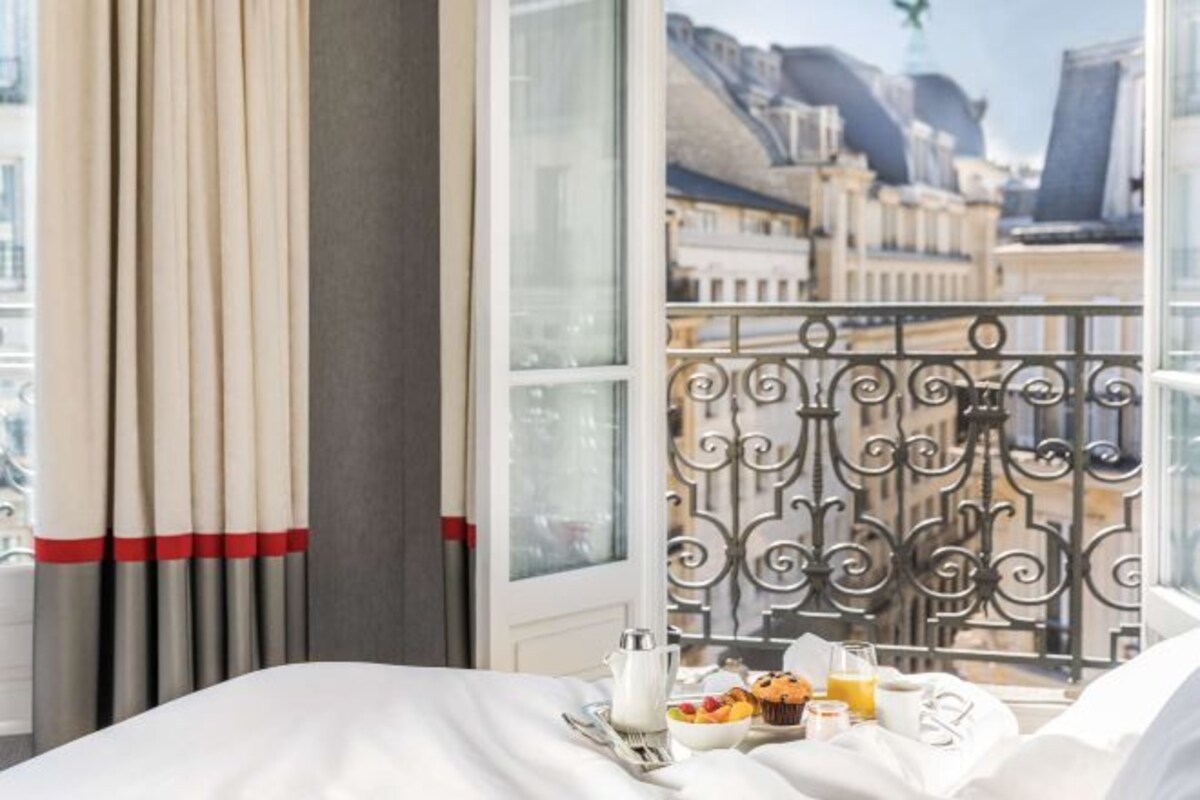 A hotel room with a bed, a tray with breakfast items, a window with a view of buildings, and curtains.