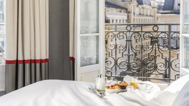 A hotel room with a balcony, a bed with white linens, a tray with breakfast items, and a view of city buildings. - Maison Albar - Le Diamond (Paris)