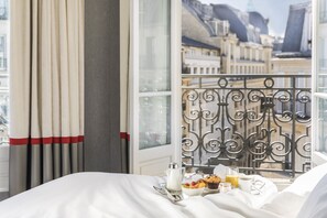 A hotel room with a balcony, a bed with white linens, a tray with breakfast items, and a view of city buildings. - Maison Albar - Le Diamond (Paris)