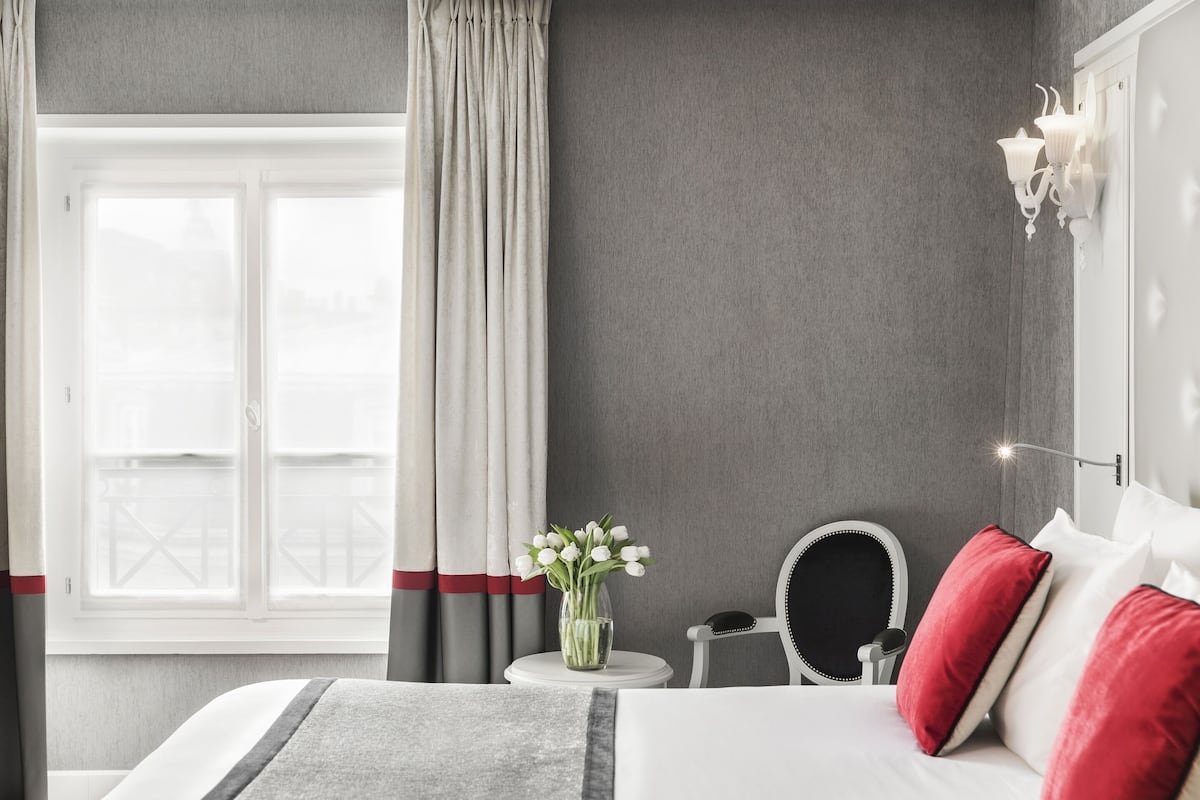 A hotel room with a bed, a round mirror, a small table with a vase of flowers, and a window with curtains.