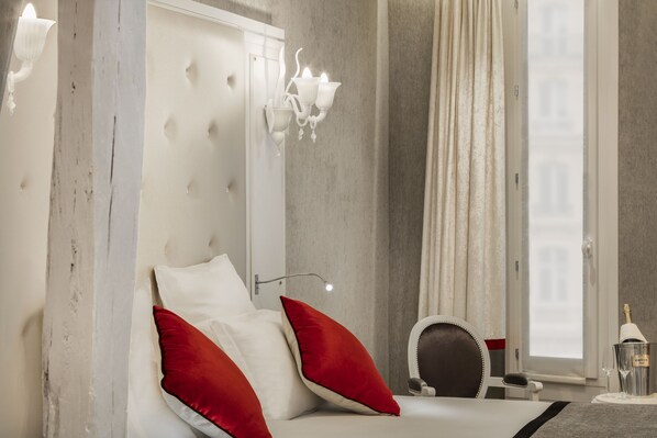 A modern hotel room with a large bed, red and white pillows, a white headboard, a wall-mounted light fixture, a mirror, and a window with curtains. - Maison Albar - Le Diamond (Paris)