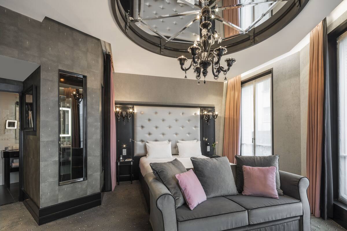 A modern hotel room with a large bed, a sofa, a chandelier, and a view of the city.