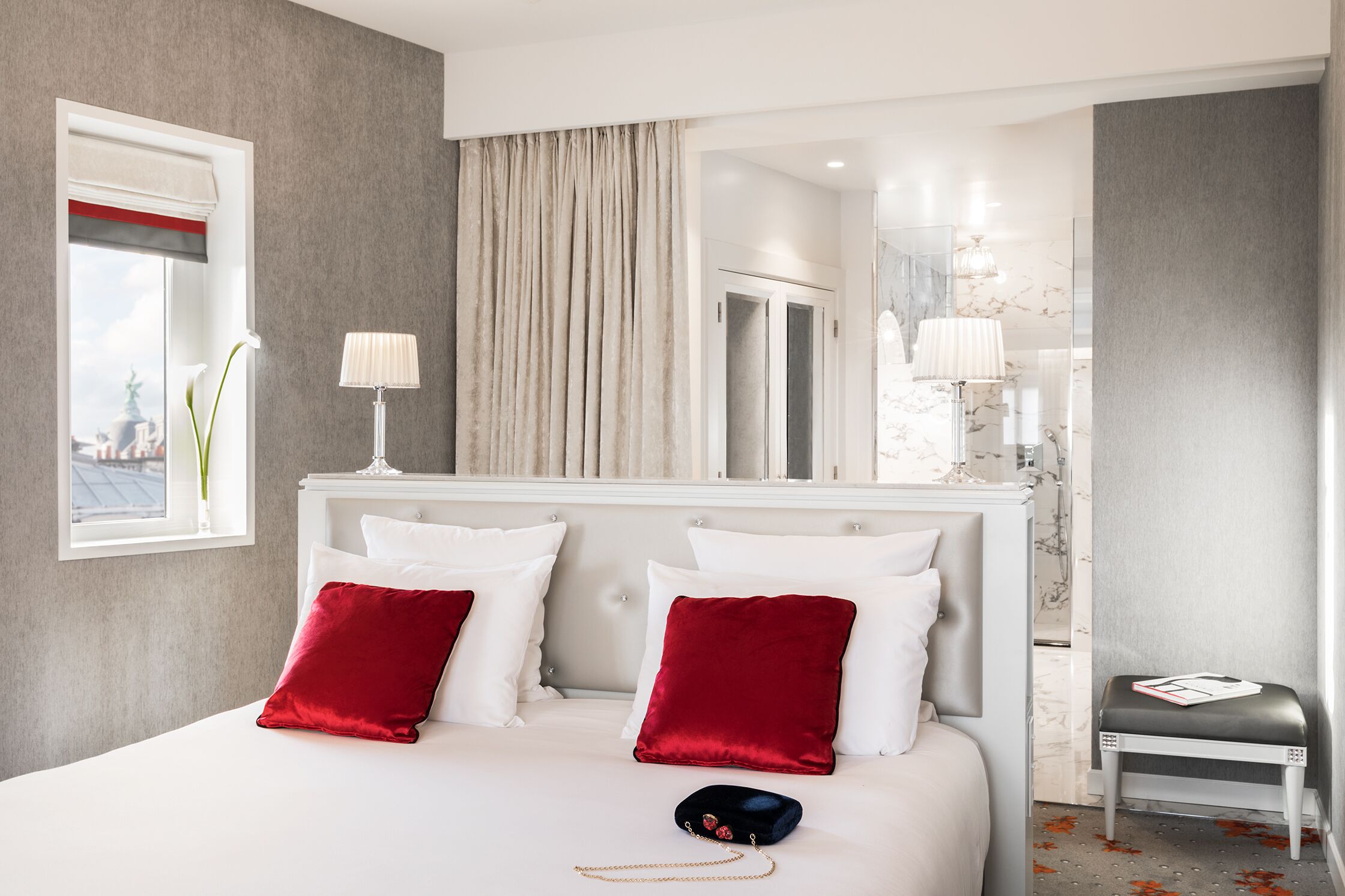 Ein Hotelzimmer mit einem großen Bett, zwei roten Kissen, einem Nachttisch mit Lampe, einem kleinen Hocker und Blick auf die Stadt durch ein Fenster mit rotem und weißem Vorhang.