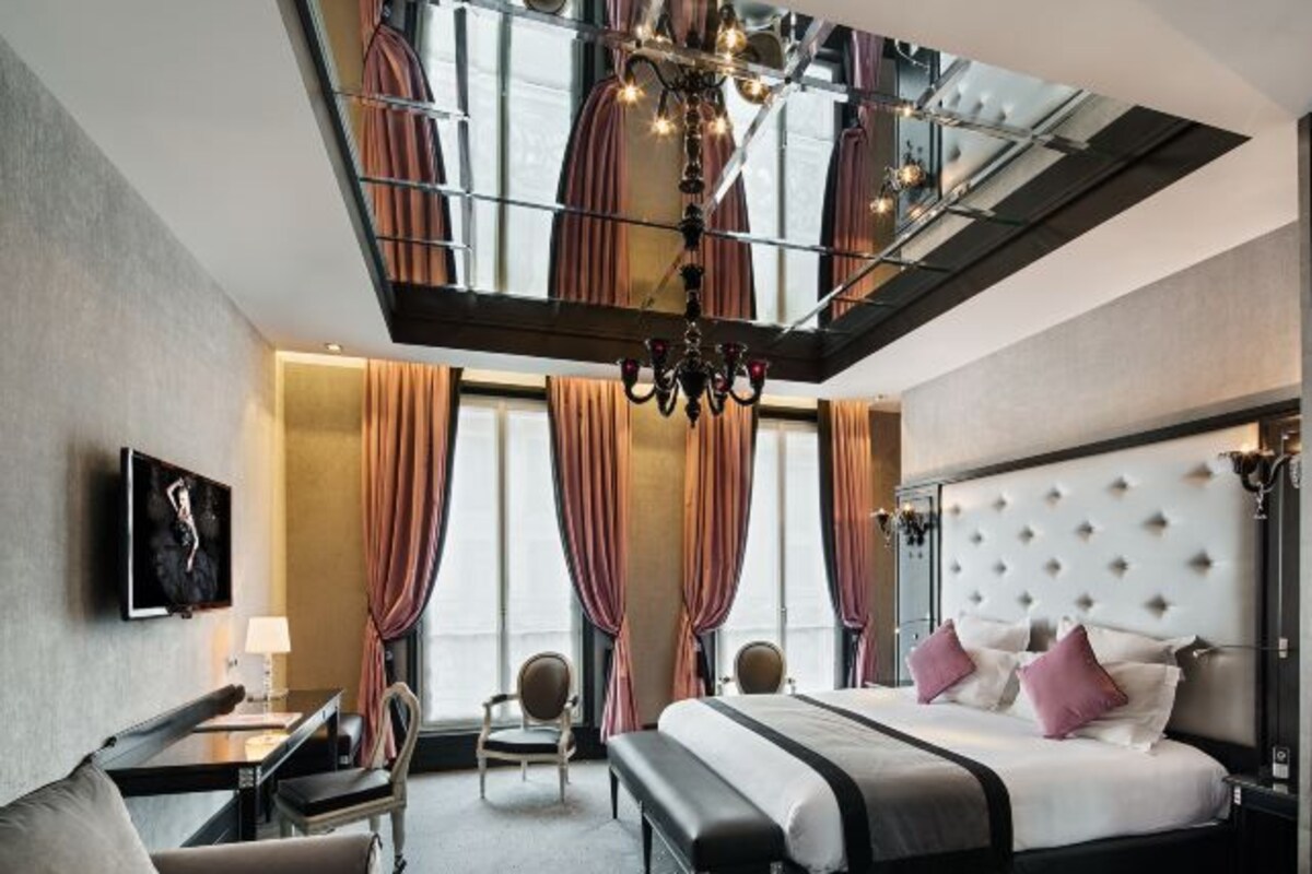 A modern hotel room with a large bed, a desk, and a chair. There is a chandelier hanging from the ceiling and curtains on the windows.