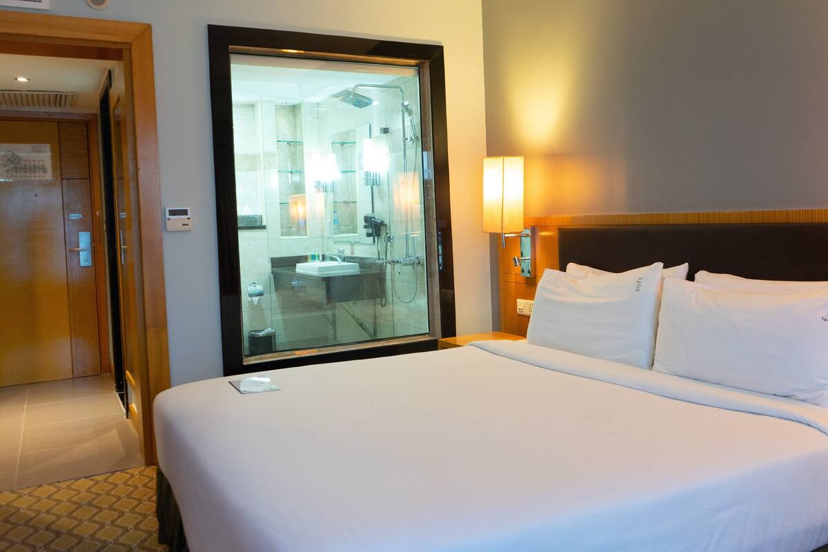 A hotel room with a bed, a large mirror, a bathroom visible through the mirror, and a nightstand with a lamp.