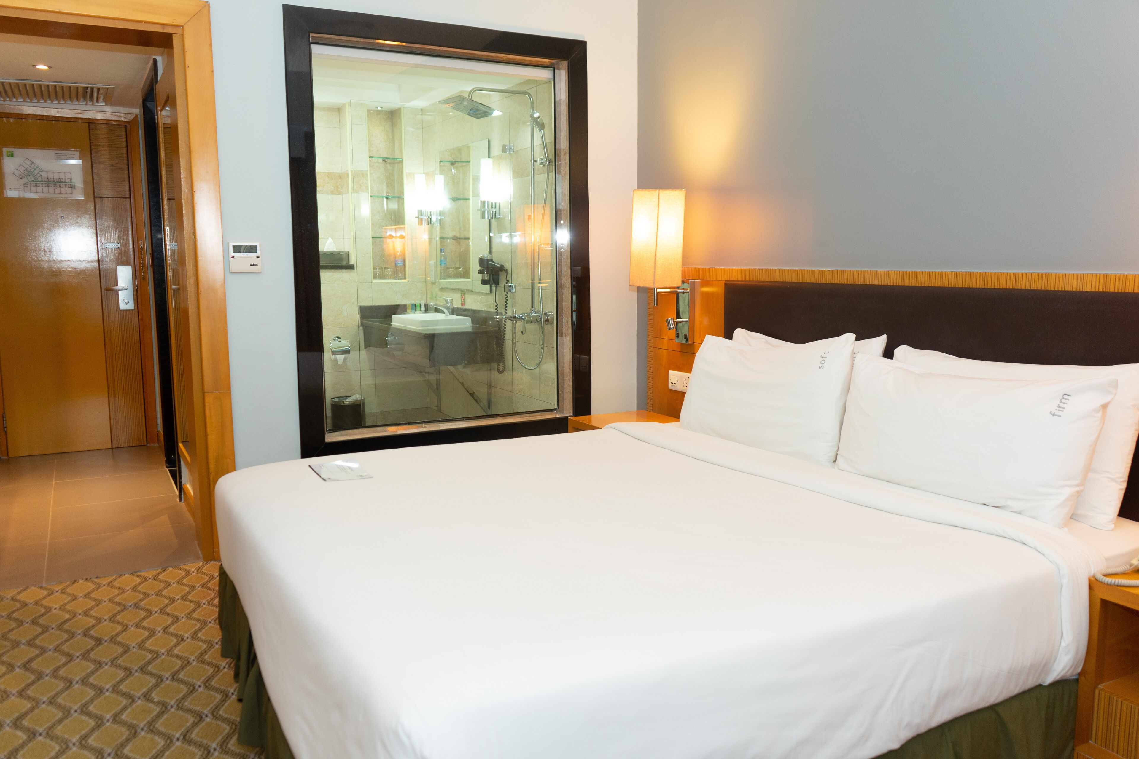A hotel room with a large bed, a bedside table, a nightstand, a mirror, a window, and a bathroom visible through the glass door.