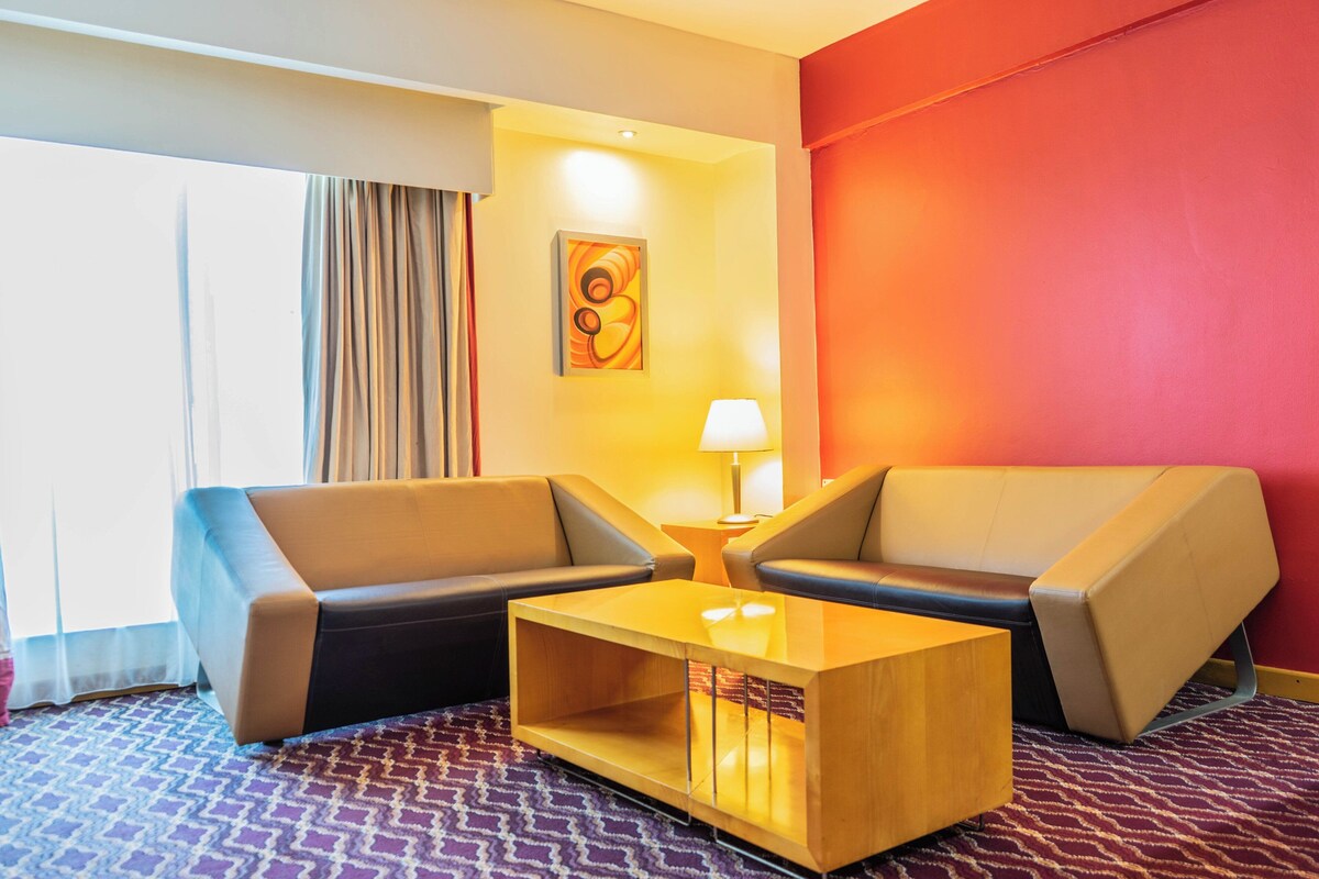 Habitación de hotel con dos sofás, una mesita de centro, una lámpara, un cuadro y una ventana con cortinas.