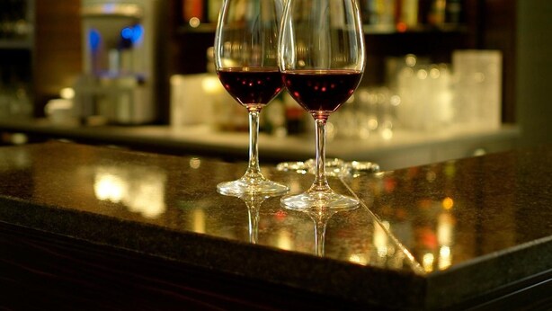 Deux verres à vin contenant un liquide rouge sont posés sur le comptoir d’un bar, avec en arrière-plan, flou, des étagères remplies de bouteilles.