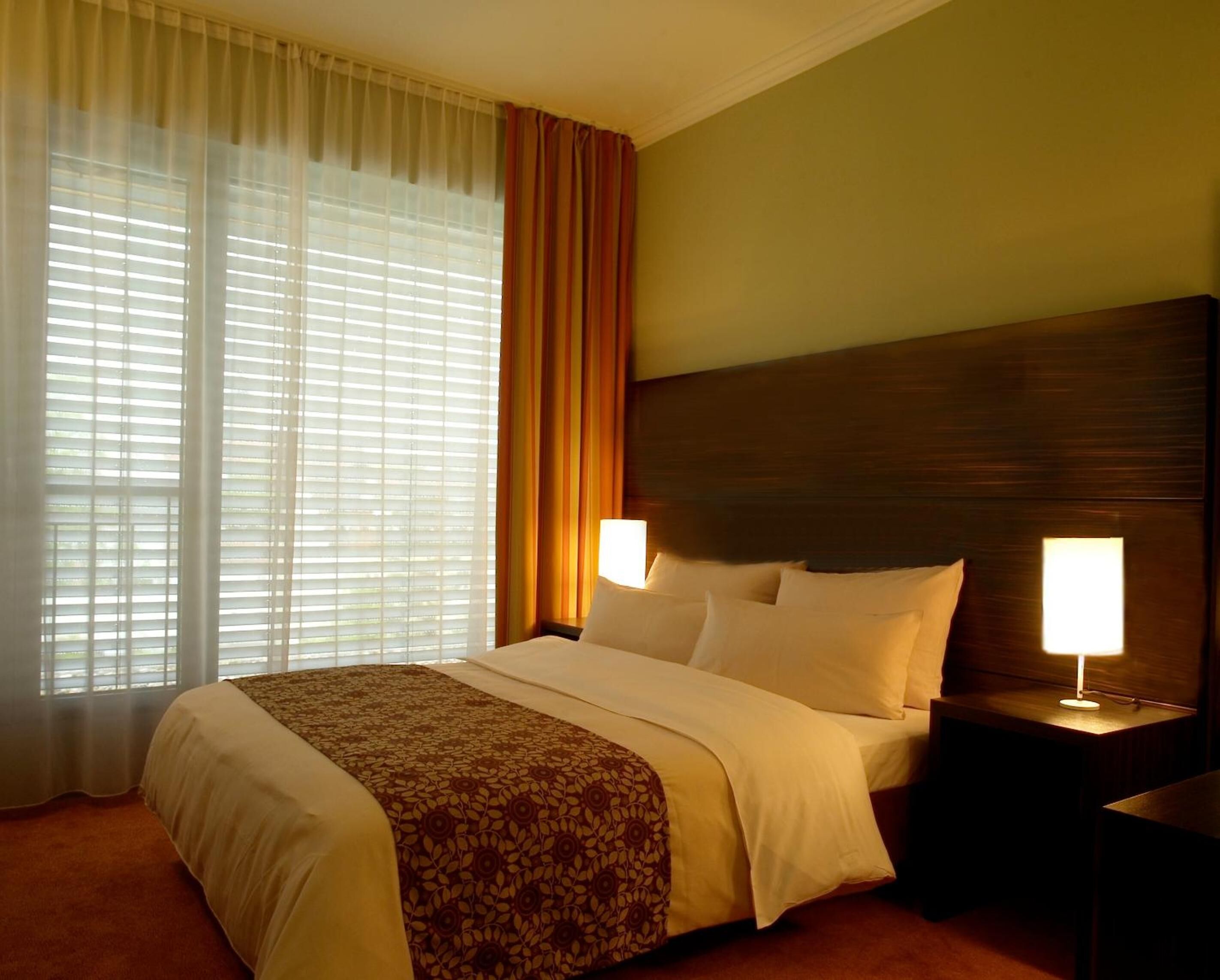 Una habitación de hotel con una cama grande, dos lámparas de noche, una ventana con persianas y una cortina.