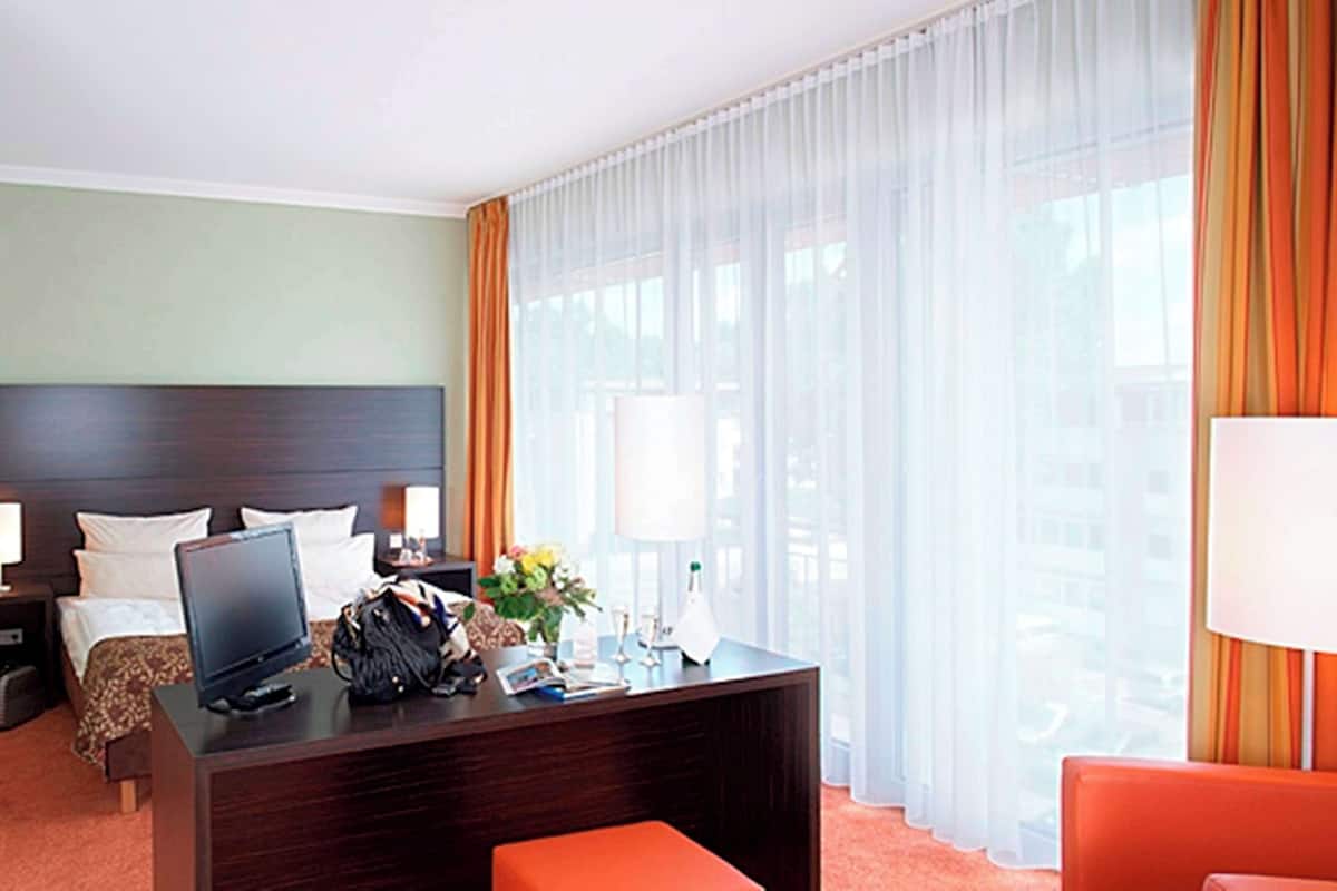 Une chambre d’hôtel avec un lit, un bureau équipé d’un ordinateur, une chaise, une lampe et une grande fenêtre avec des rideaux.