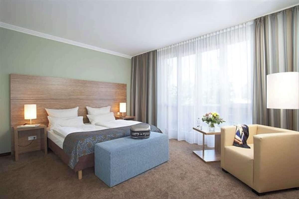 Une chambre d’hôtel avec un grand lit, deux tables de chevet avec des lampes, une petite table d’appoint avec un vase, un canapé et une fenêtre avec des rideaux.