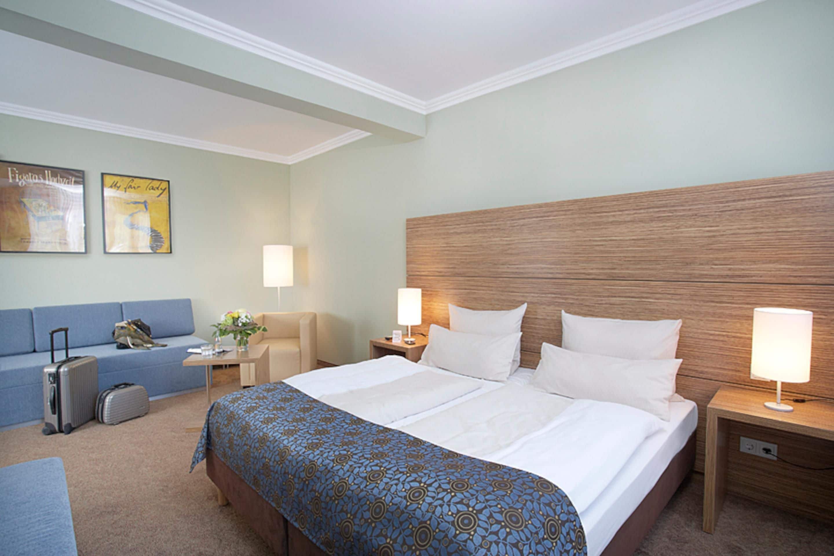 Ein Hotelzimmer mit einem großen Bett, einem blauen Sofa, einem Schreibtisch mit Lampe und einem Koffer.