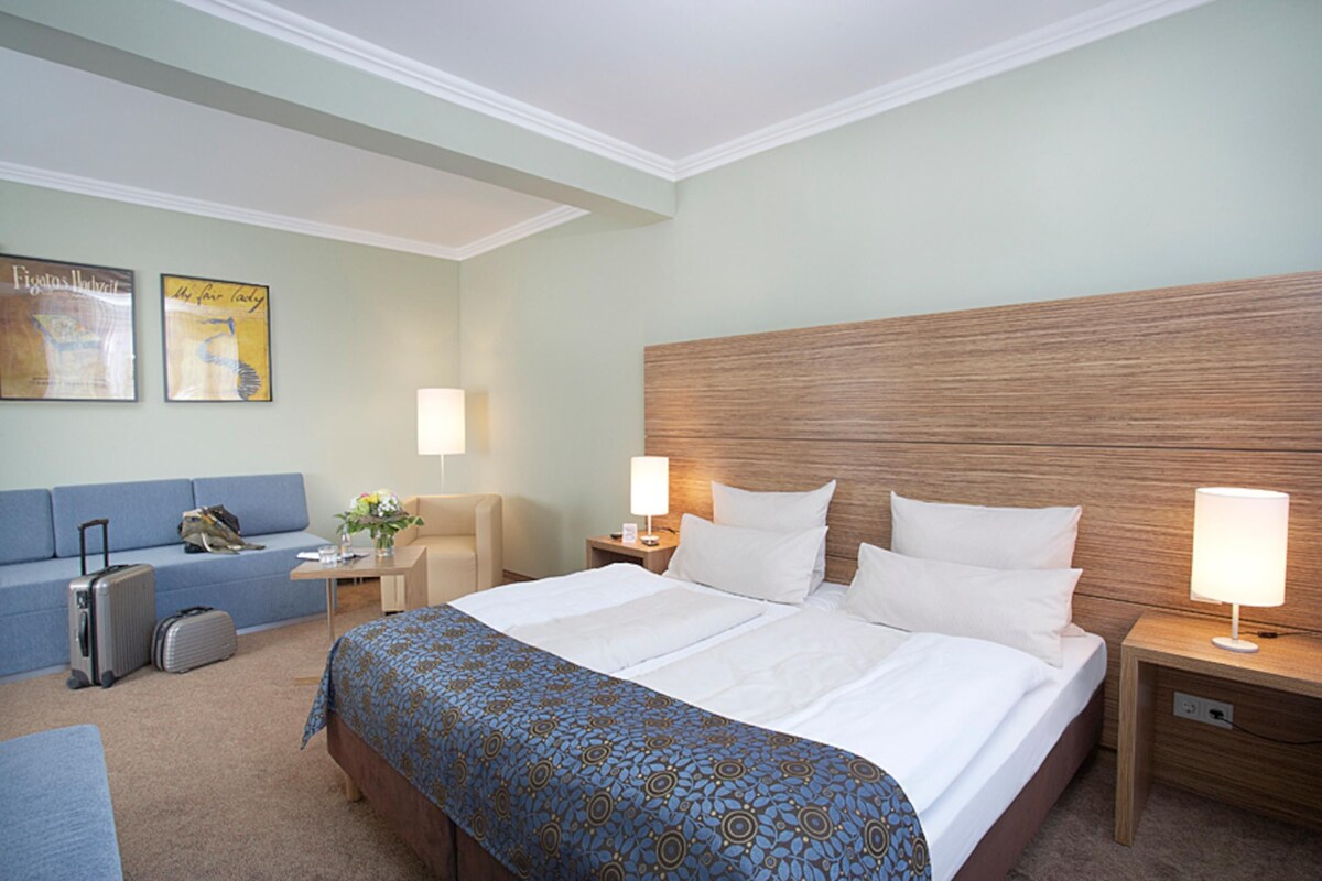 Une chambre d’hôtel avec un grand lit, un canapé bleu, un bureau avec une lampe et une valise.