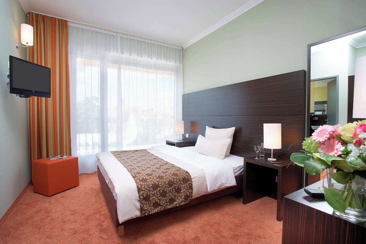 Une chambre d’hôtel comprenant un lit, une télévision, une table de chevet avec une lampe, une commode et un vase avec des fleurs.