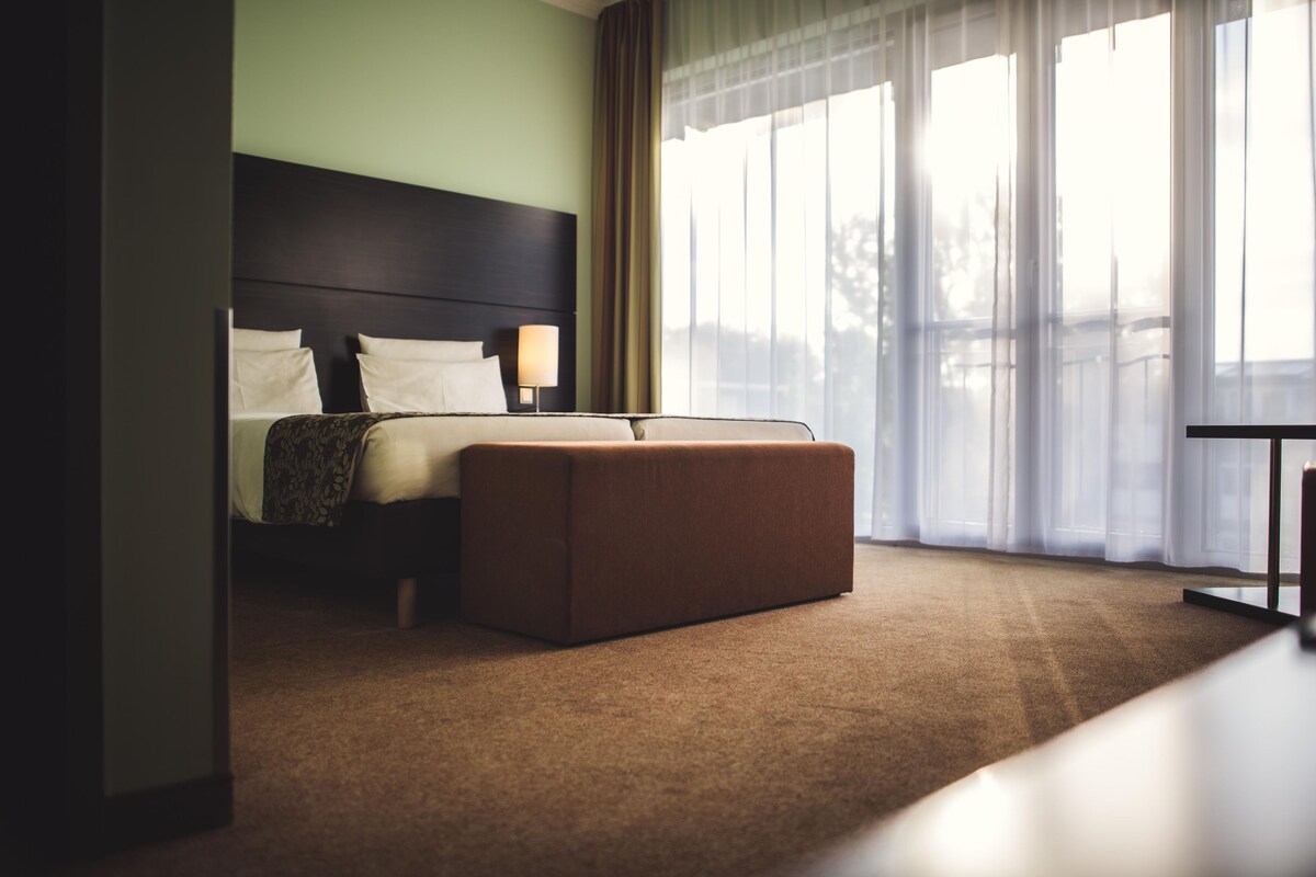 Une chambre d’hôtel avec un lit, un pouf, une table de chevet et une fenêtre avec des rideaux.