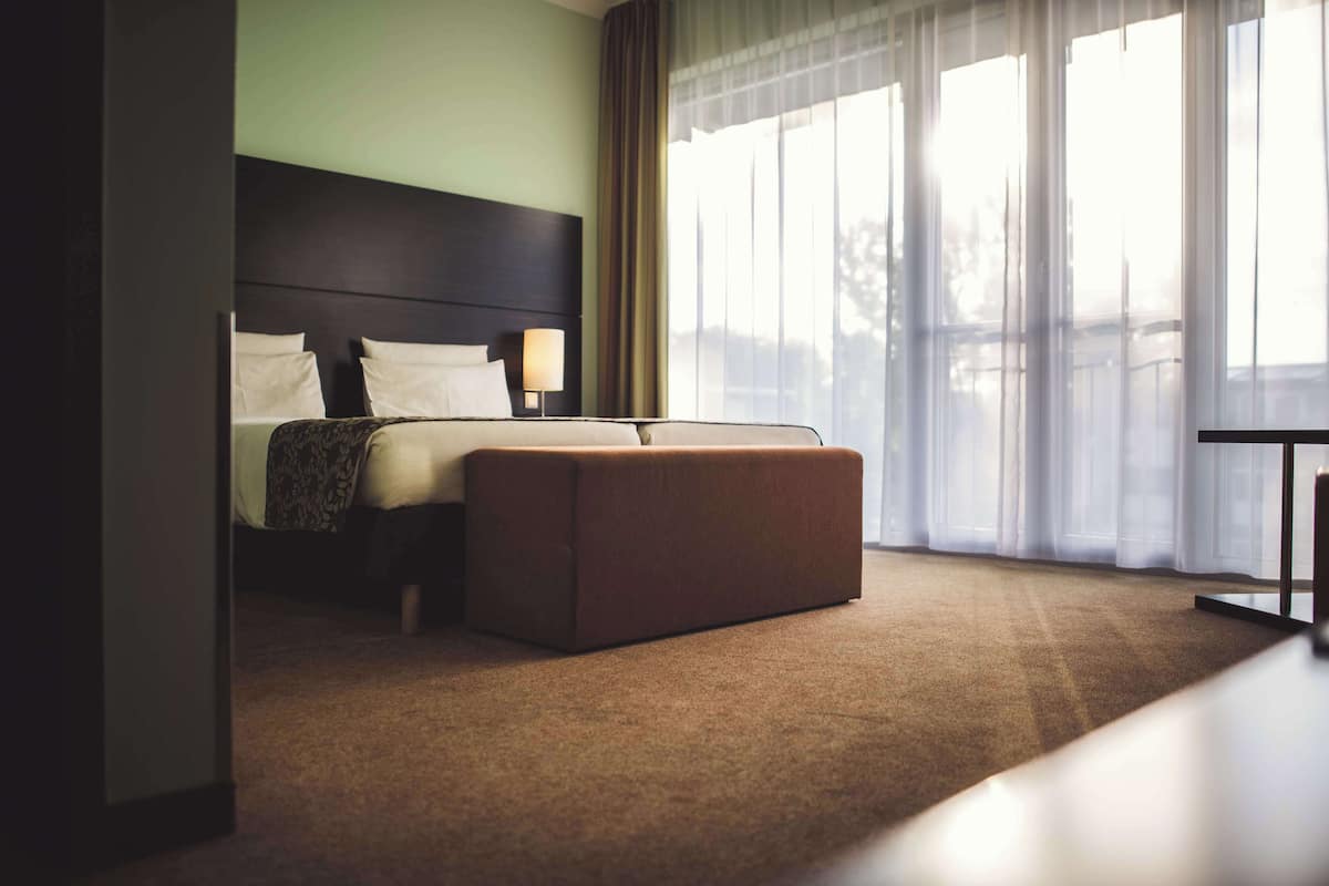 Une chambre d’hôtel avec un lit, une table de chevet, une lampe, une grande fenêtre avec des rideaux légers et une vue sur l’extérieur.