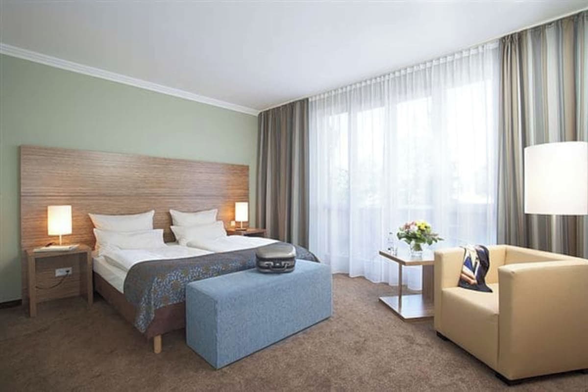 Una habitación de hotel con una cama grande, dos mesitas de noche con lámparas, una mesita auxiliar con un jarrón, un sofá y una ventana con cortinas.