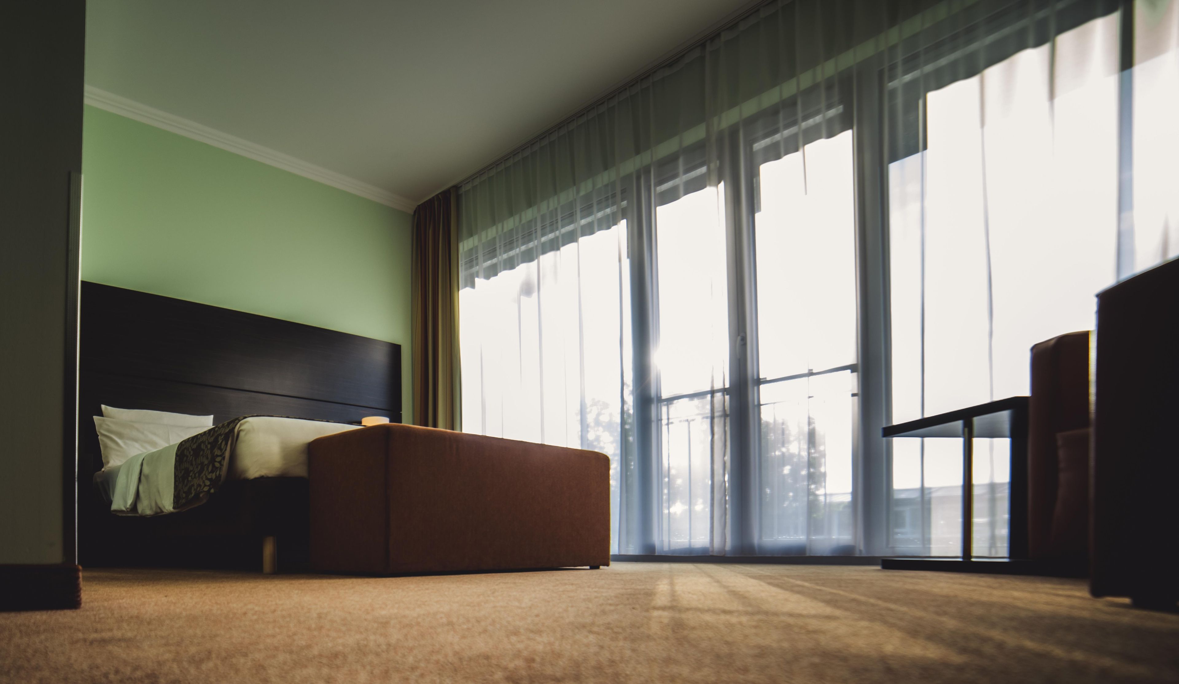 A hotel room with a large bed, a brown ottoman, and a view of the city through large windows.