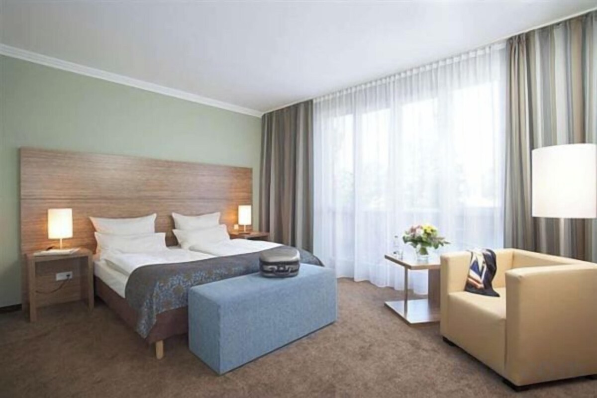 A hotel room with a bed, bedside tables, a lamp, a sofa, and a window with curtains.