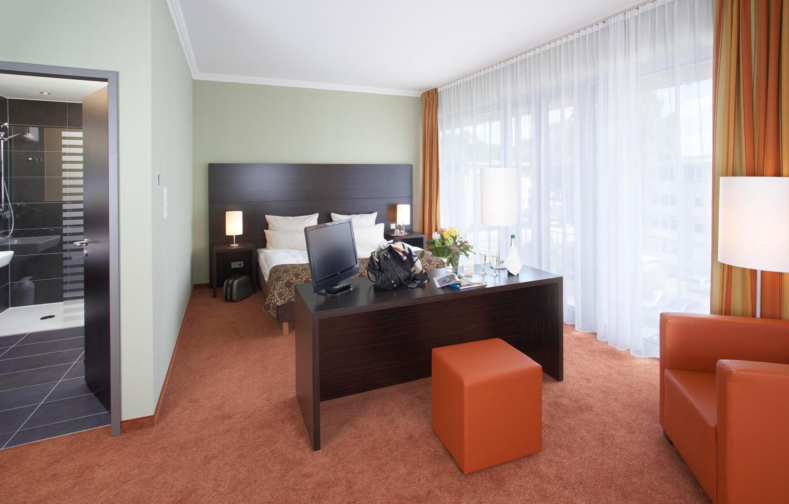 A hotel room with a bed, desk, and chair, a bathroom with a shower, and a view of the city through large windows.
